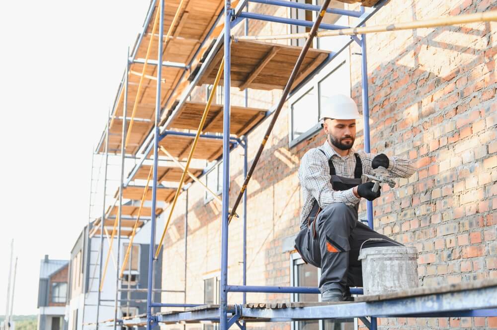 Construction worker on scaffolding applying mortar with trowel to brick exterior wall.